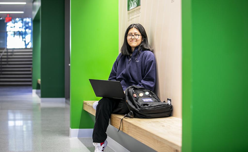 A photo of an undergraduate student sitting on a bench in a hallway with a backpack and a laptop.